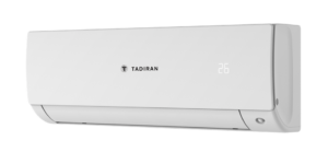 יחידת פנים עילית מעוצבת של מזגן Tadiran Mini VRF בגוון לבן נקי, מותקנת בצורה גלויה על קיר מעוצב בסלון מודרני או במשרד. לצד היחידה מוצג השלט הרחוק / התרמוסטט מדגם PRIME-HQS01. היחידה מציגה קווים נקיים, אסתטיים וזרימת אויר שקטה.